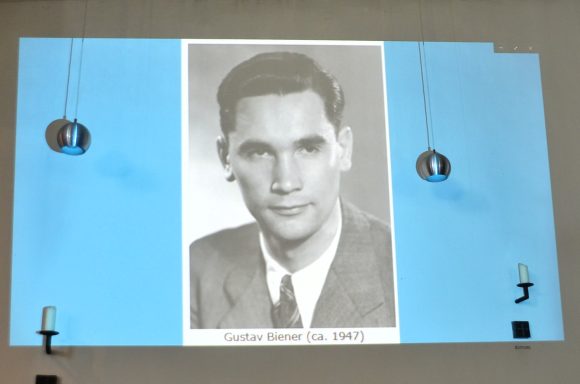 Gustav Biener Porträt eines Mannes in Anzug, projiziert auf eine blaue Wand.